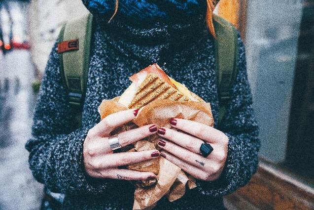 Person holding a large sandwich wrapped in brown paper, wearing a grey sweater and green backpack. Person holding a large sandwich wrapped in brown paper, wearing a grey sweater and green backpack.