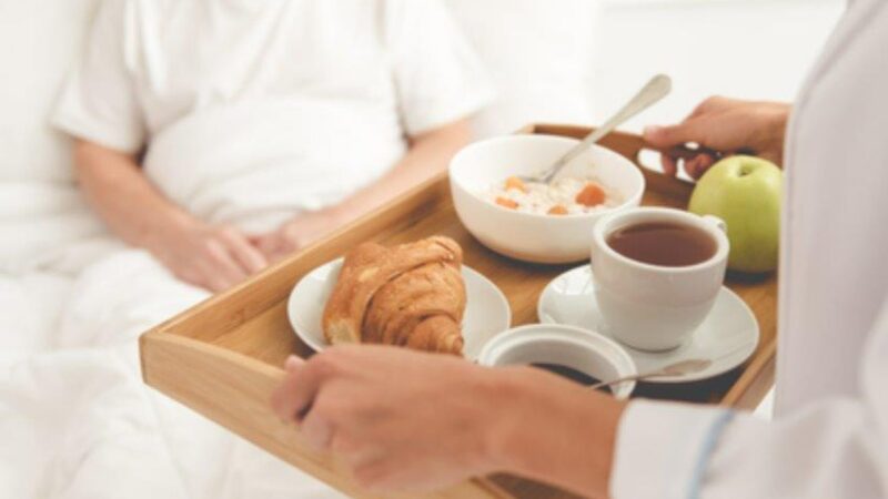 Healthcare worker serving breakfast on a wooden tray to a patient in bed, including a croissant, tea, apple, and a bowl of cereal Healthcare worker serving breakfast on a wooden tray to a patient in bed, including a croissant, tea, apple, and a bowl of cereal