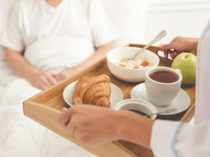 Healthcare worker serving breakfast on a wooden tray to a patient in bed, including a croissant, tea, apple, and a bowl of cereal
