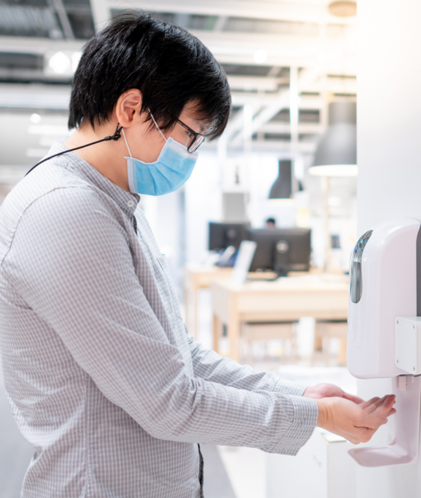 A person in a mask standing in front of a hand sanitiser dispenser A person in a mask standing in front of a hand sanitiser dispenser